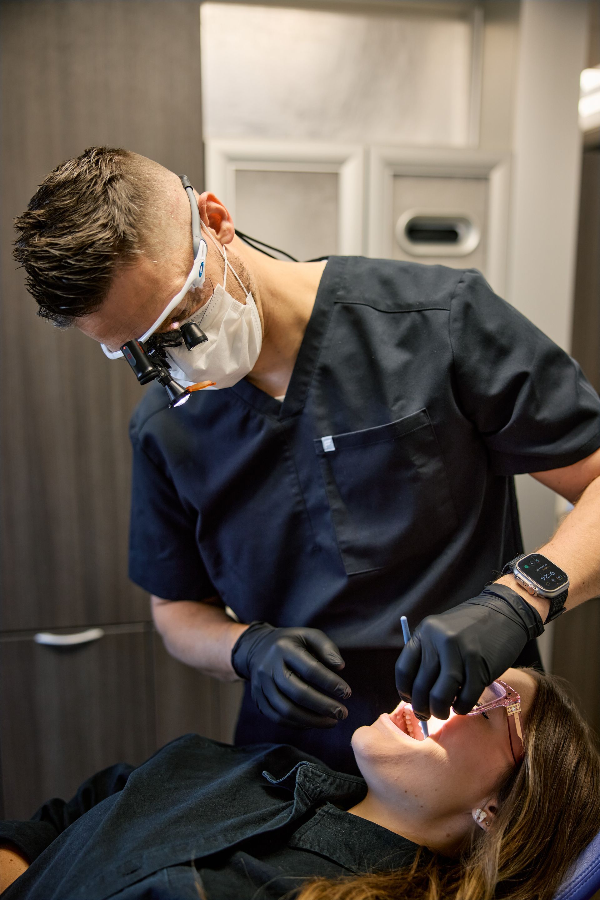 Dentist examining patient's teeth. Dentist wearing mask, loupes, and gloves. Patient is in a dental chair.