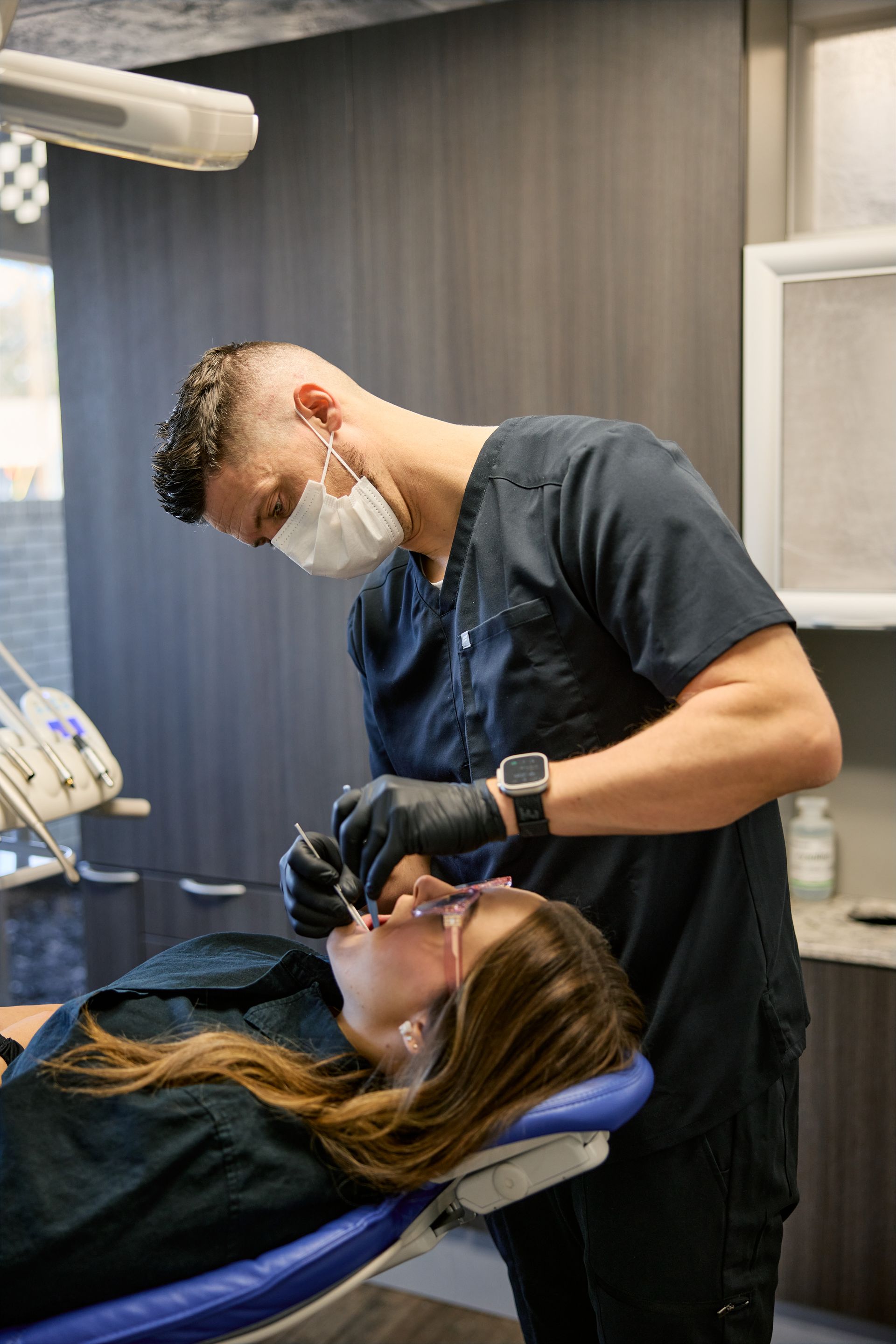 Dentist in scrubs, mask, and gloves examining a patient's teeth in a dental office.