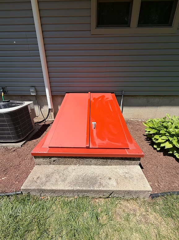 Red metal utility hatch beside a house on a concrete pad, with grass and mulch around it.