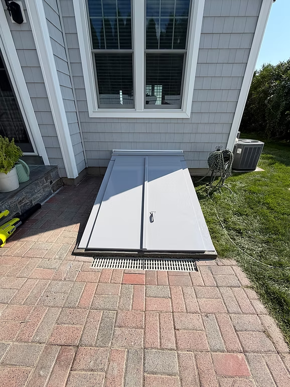 Closed gray basement bulkhead doors beside a white house, next to a brick patio and lawn.