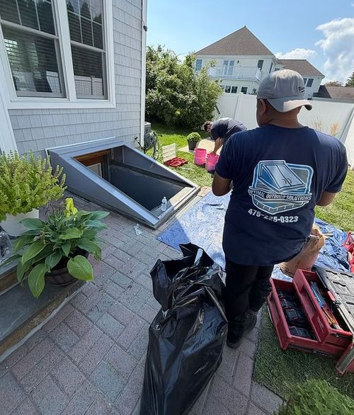 Two workers clearing debris beside a house, with black trash bags and tools on a driveway.