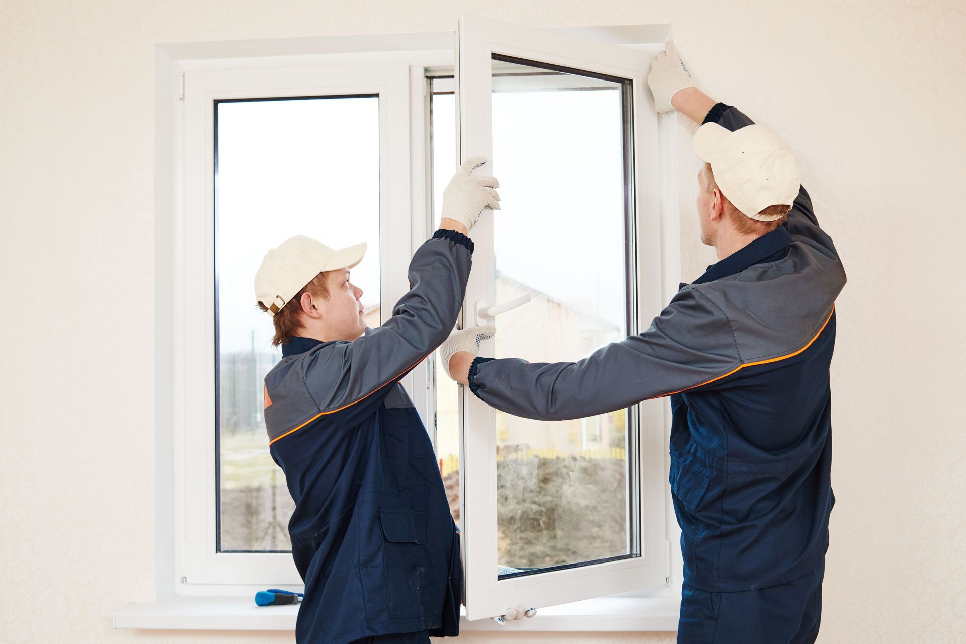 Two workers installing a white window in a room. They wear work clothes and caps, holding the frame.