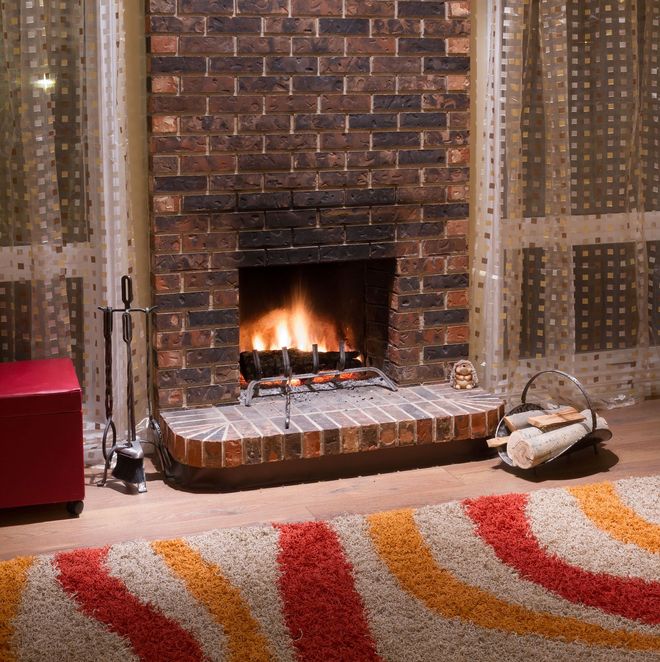 Fireplace with burning fire, brick surround, logs, and a colorful rug in a living room.