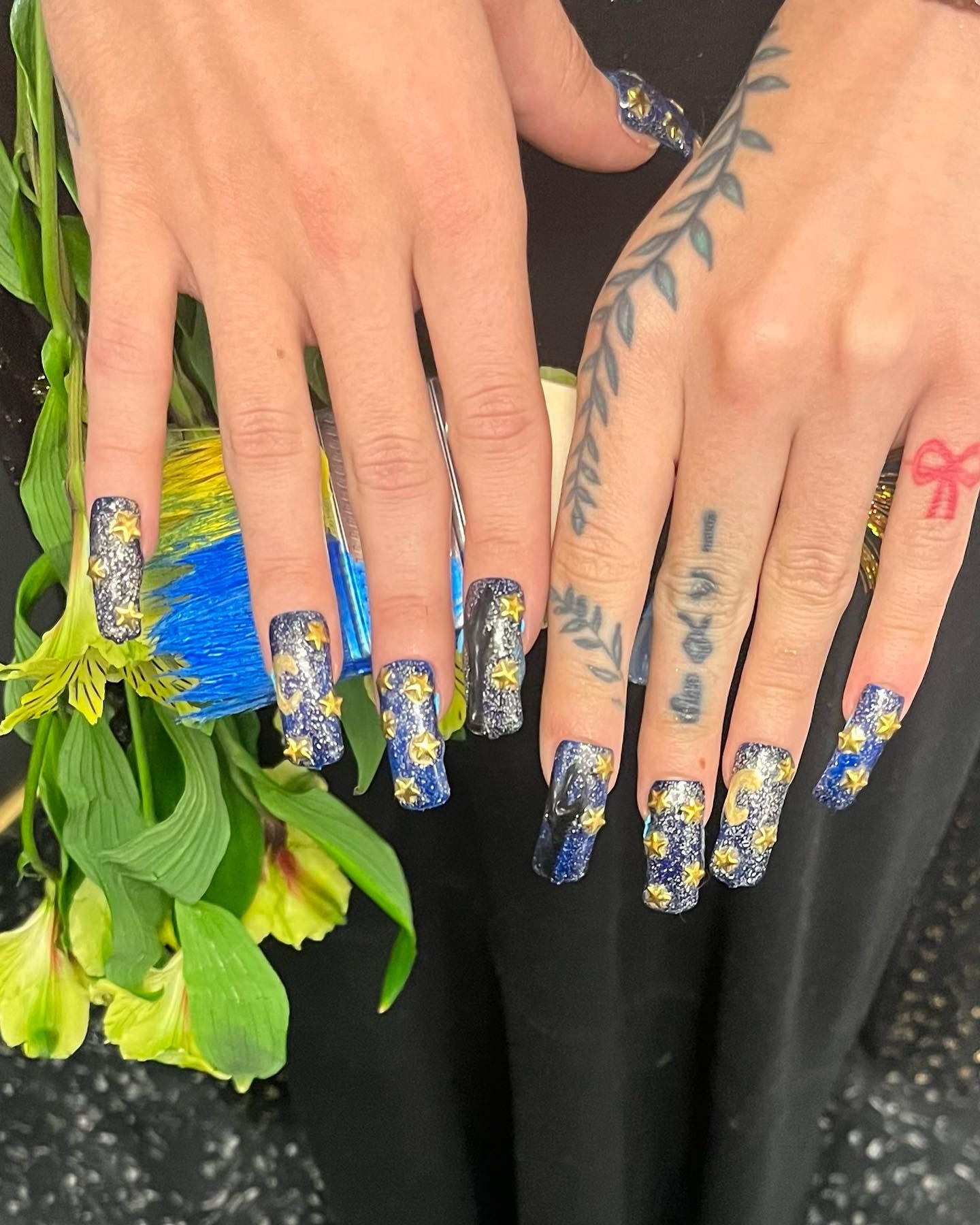 Hands with long, glittery, blue and yellow daisy-decorated nails holding a flower with green leaves.