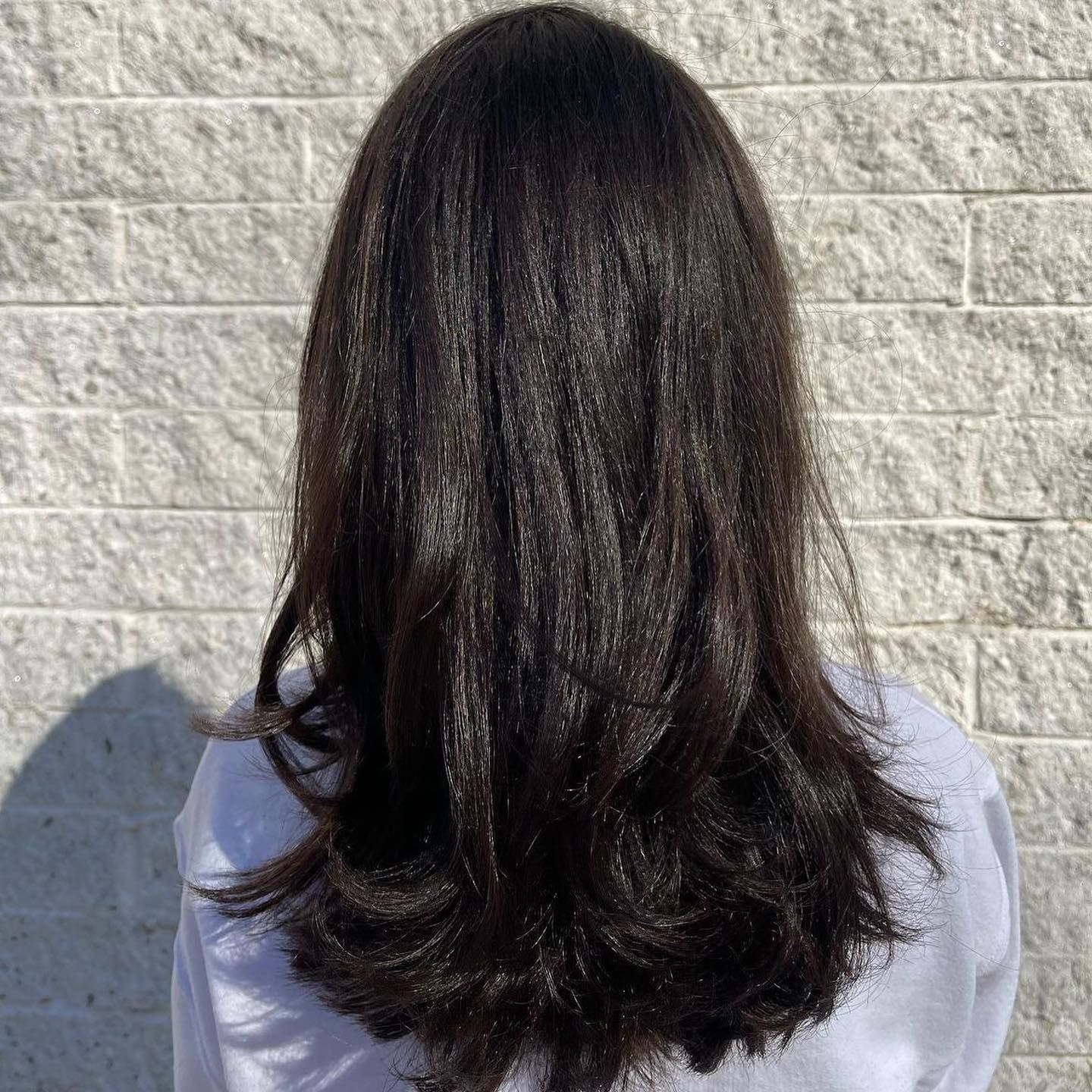 Woman with long, dark hair; the hair is styled straight with layers, against a light brick wall.