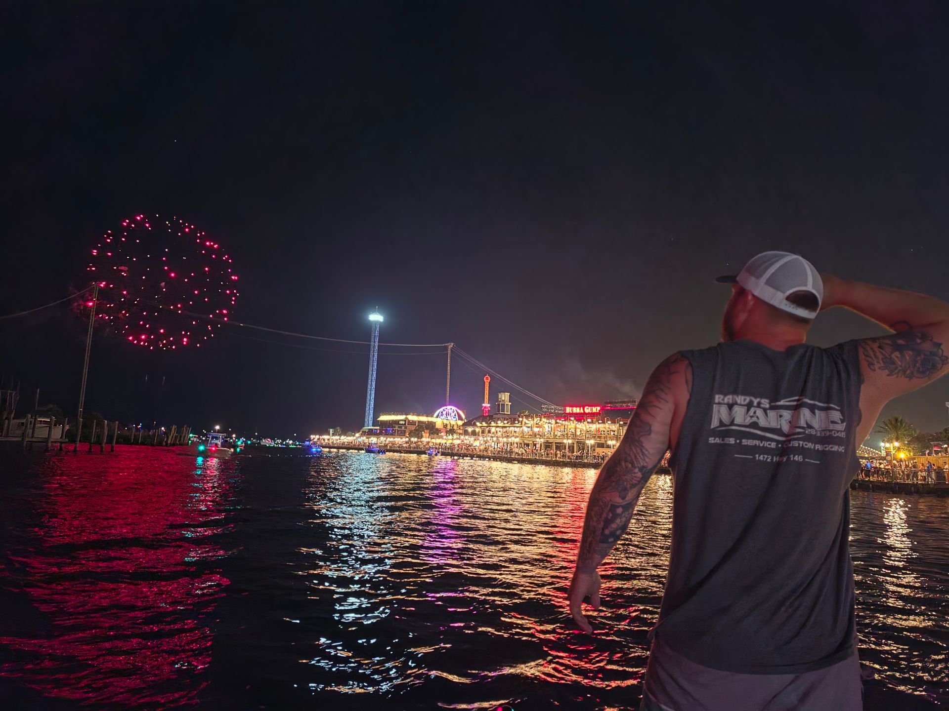 Person in a sleeveless shirt watches fireworks over water at night, with city lights reflecting on the surface.