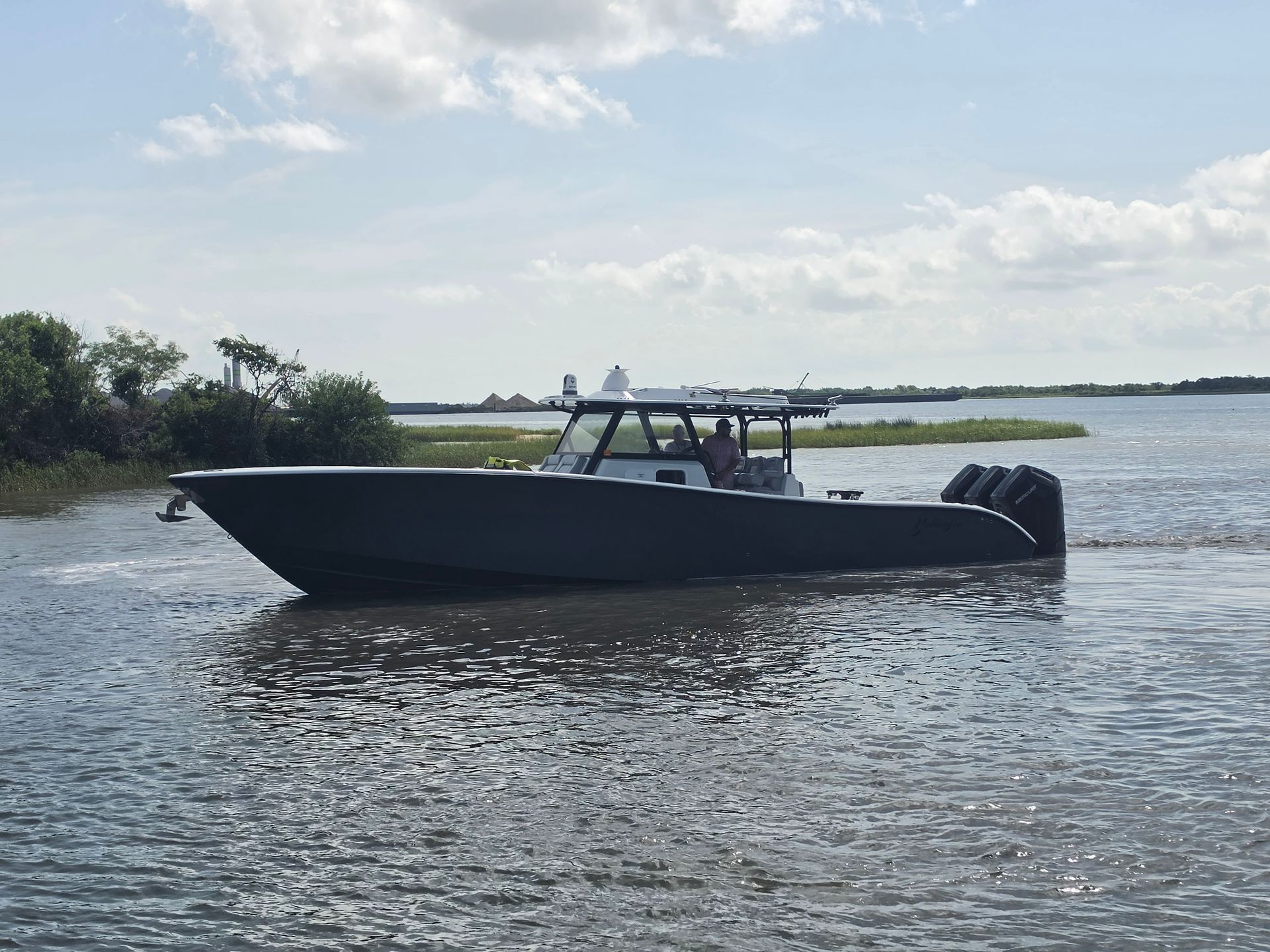Dark motorboat on calm water near a grassy shoreline under a partly cloudy sky