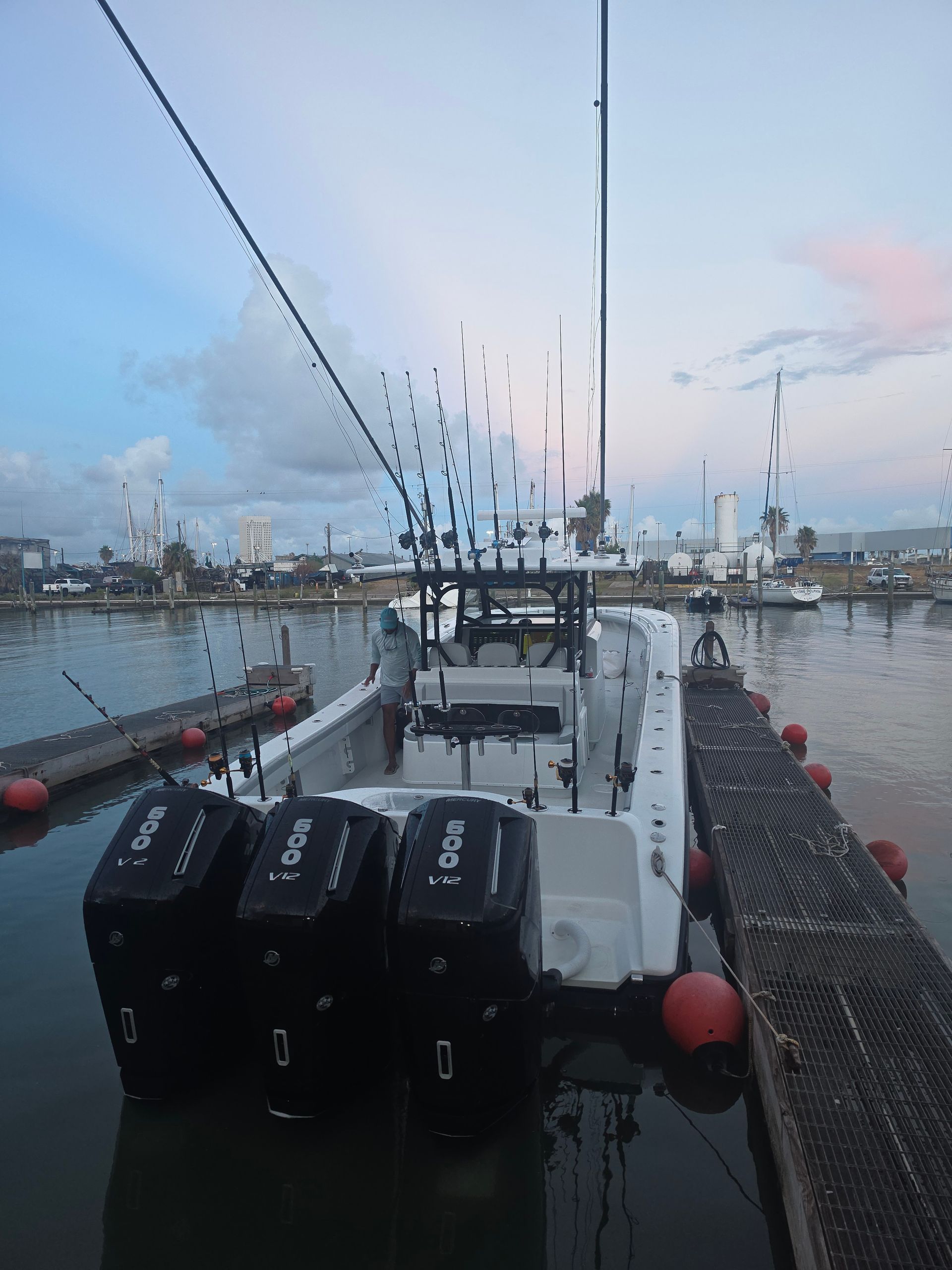 Fishing boat tied at a harbor dock at dusk, with calm water and city skyline in the background.