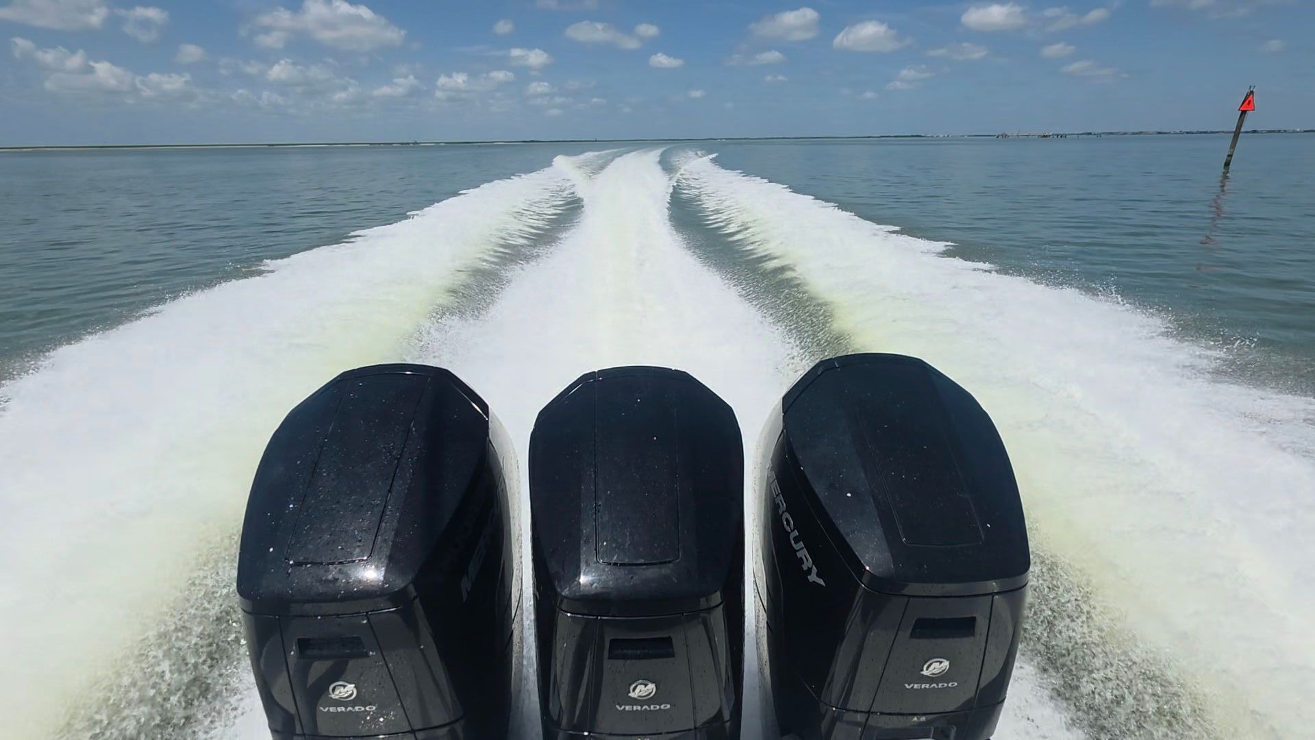 View from a speedboat with three outboard motors leaving a white wake on calm blue water under a cloudy sky