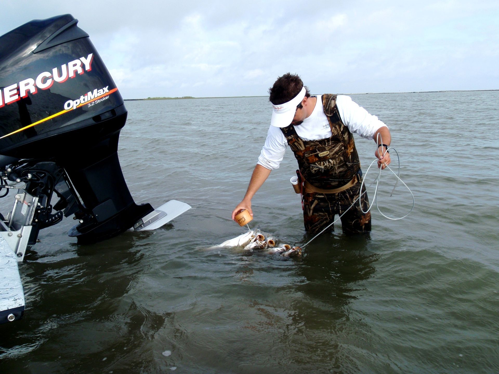 Man in waders releasing a fish into the water, near a boat with a Mercury engine. Overcast day.
