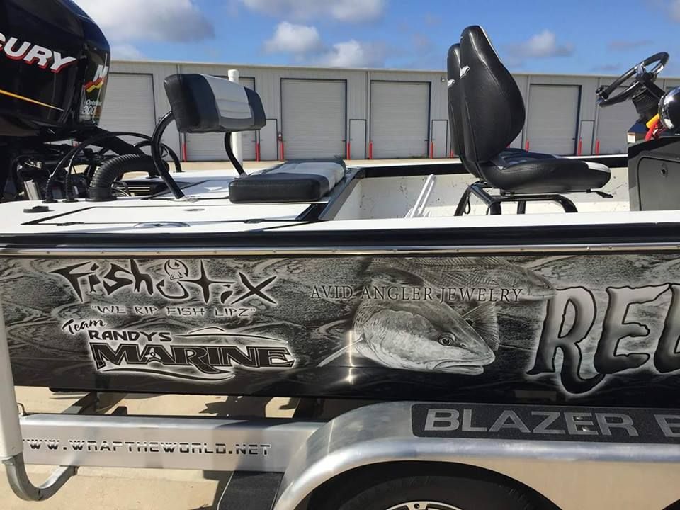 A fishing boat with a fish graphic wrap, black seating, and a Mercury engine, on a trailer in front of a storage facility.
