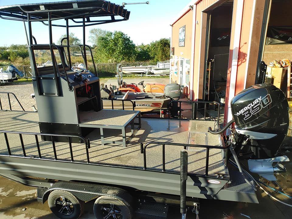 Gray fishing boat on a trailer with a Mercury outboard motor and a black tower in front of a building.