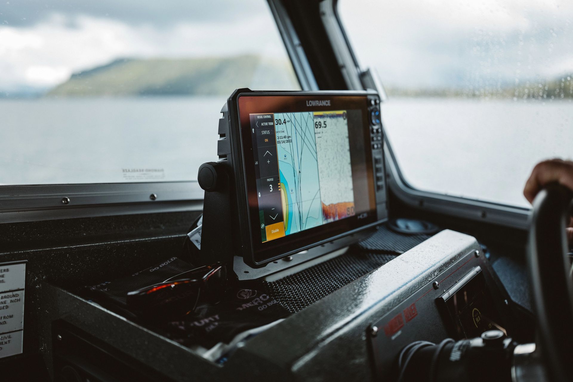 Boat dashboard with a navigation screen displaying a map; a hand on a steering wheel, water and mountains in the background.