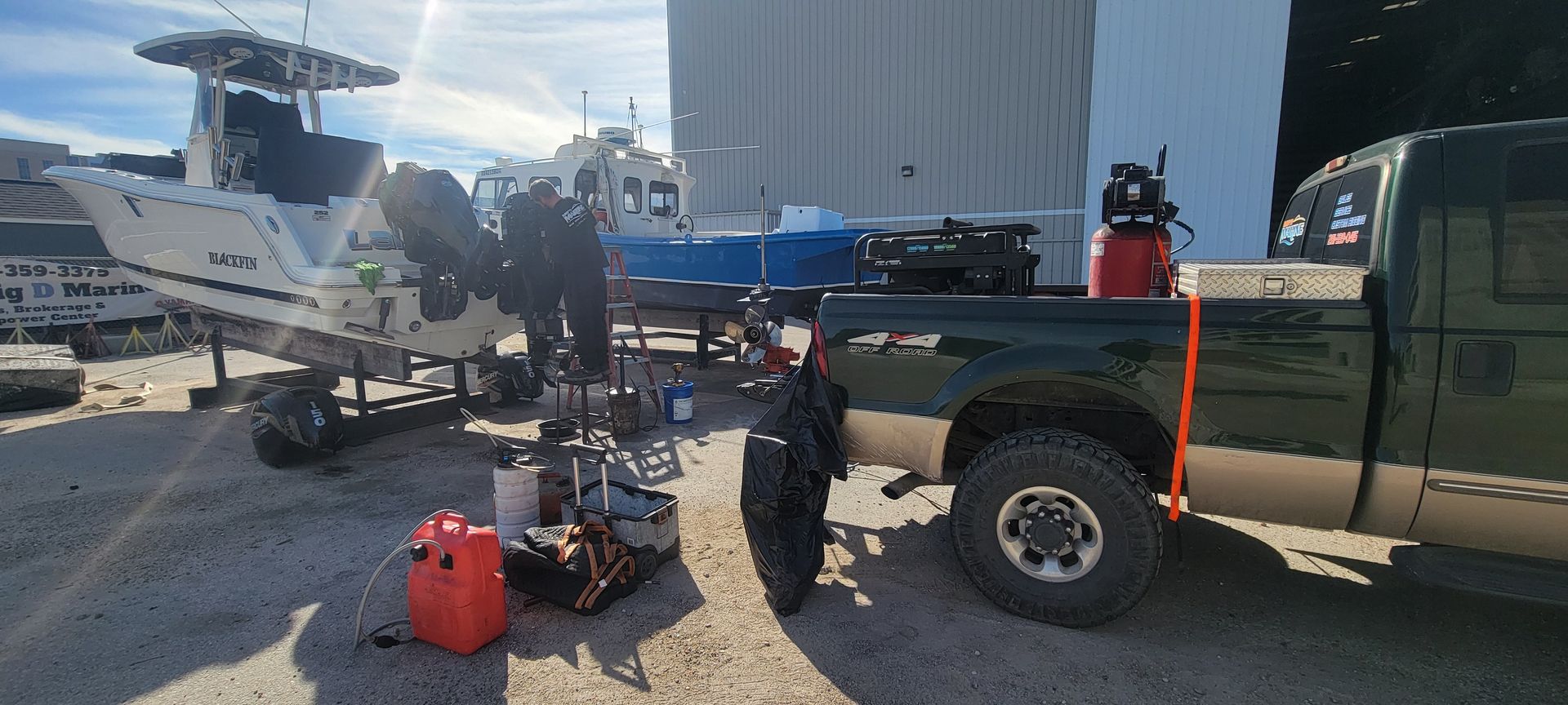 A boat on a trailer, surrounded by equipment, next to a green truck. Outdoors on a sunny day.