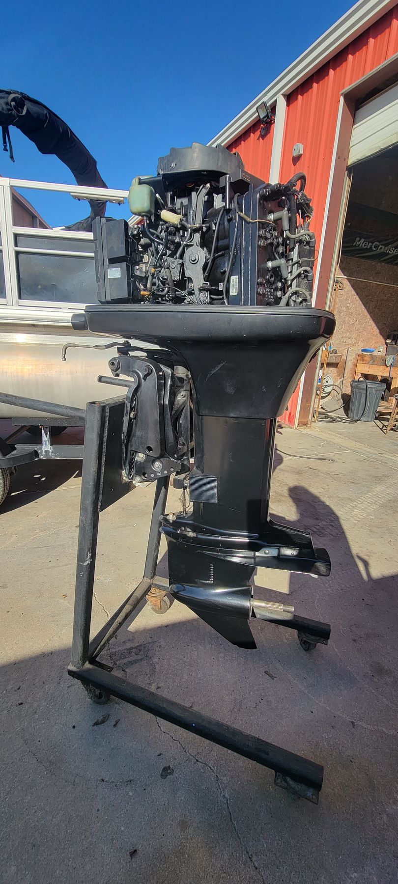 Outboard motor on a stand, black with a partially visible red building in the background, on a sunny day.
