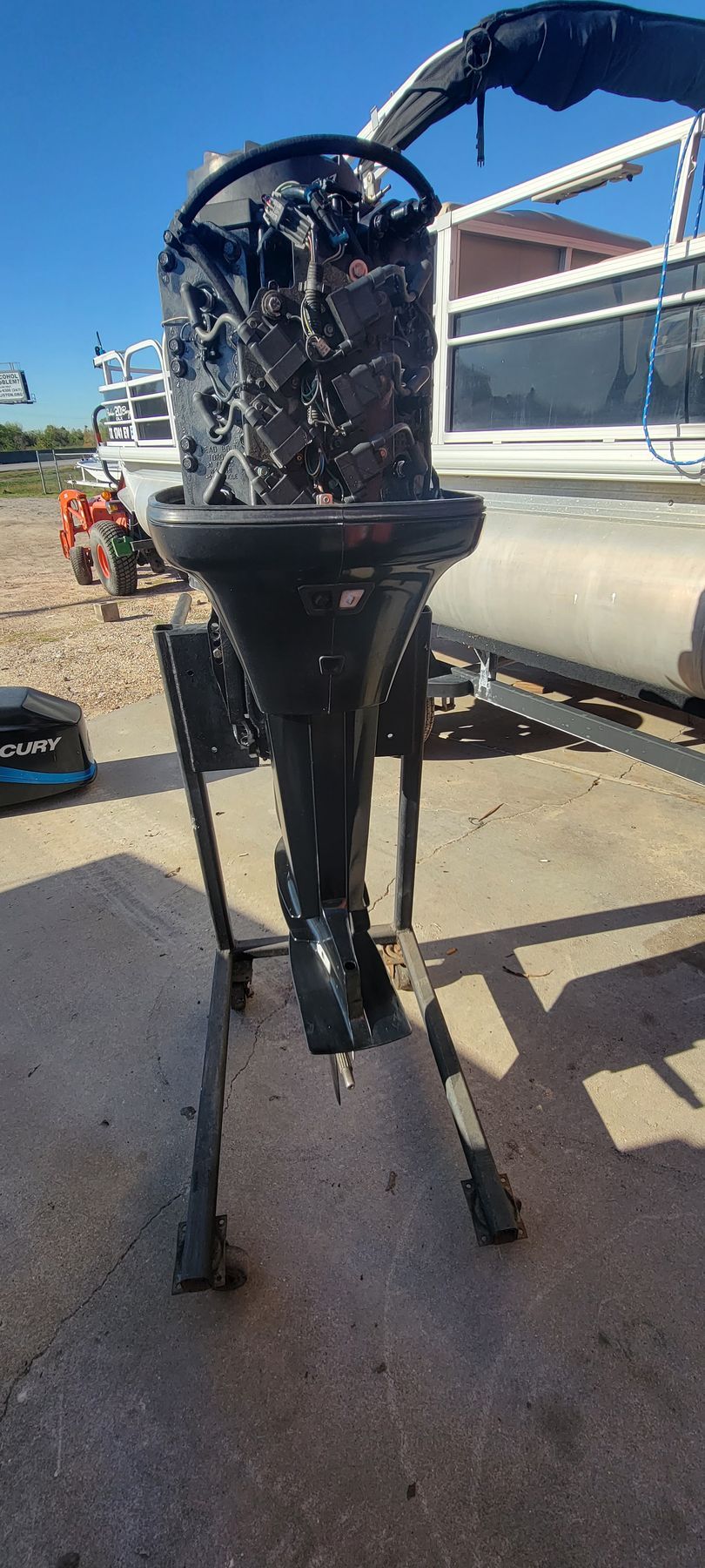 Black boat motor on a stand; pontoon boat in the background. Outdoors on concrete.