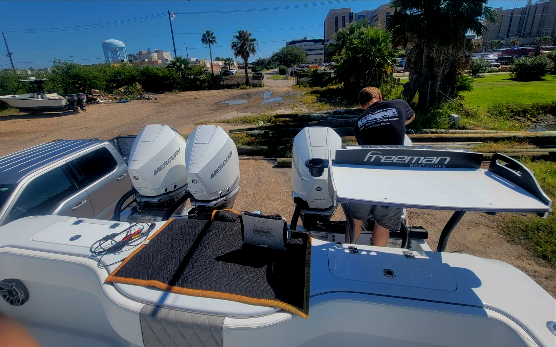 A boat with three white outboard motors parked on a dirt lot. A person stands near the motors.