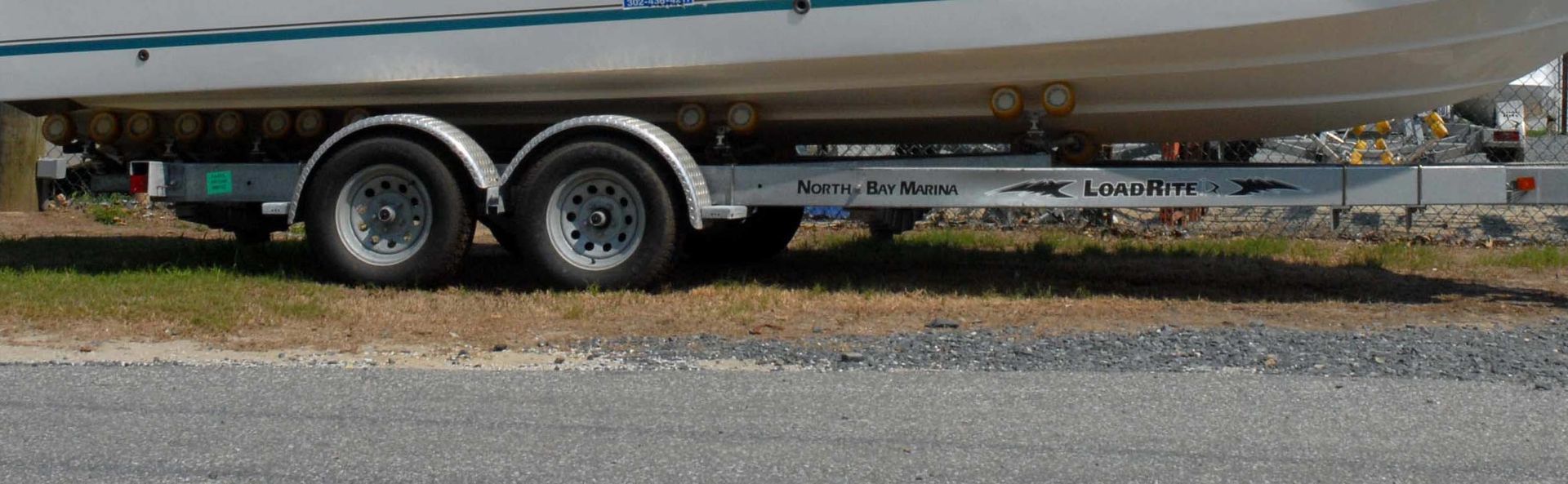 A boat trailer with two wheels on a grassy area, gray gravel road in foreground.