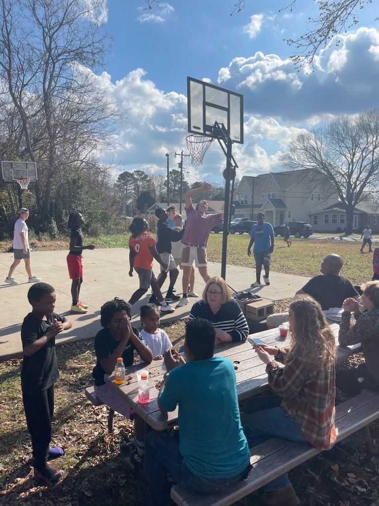 People playing basketball on a court; others seated at a picnic table. Outdoors, sunny day.