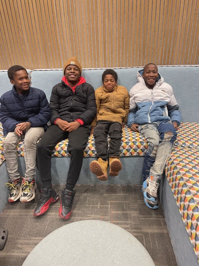 Four people smiling, sitting on a patterned couch indoors. They wear winter jackets and sneakers.