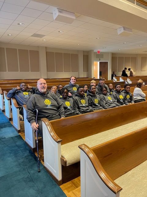 People in a church, some wearing gray hoodies, seated on pews. One man uses a crutch.