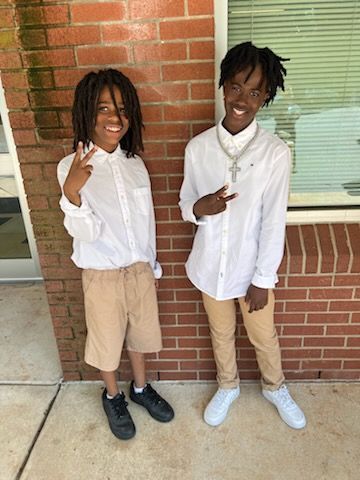 Two people in white shirts and khaki pants pose in front of a brick wall. One shows a peace sign.