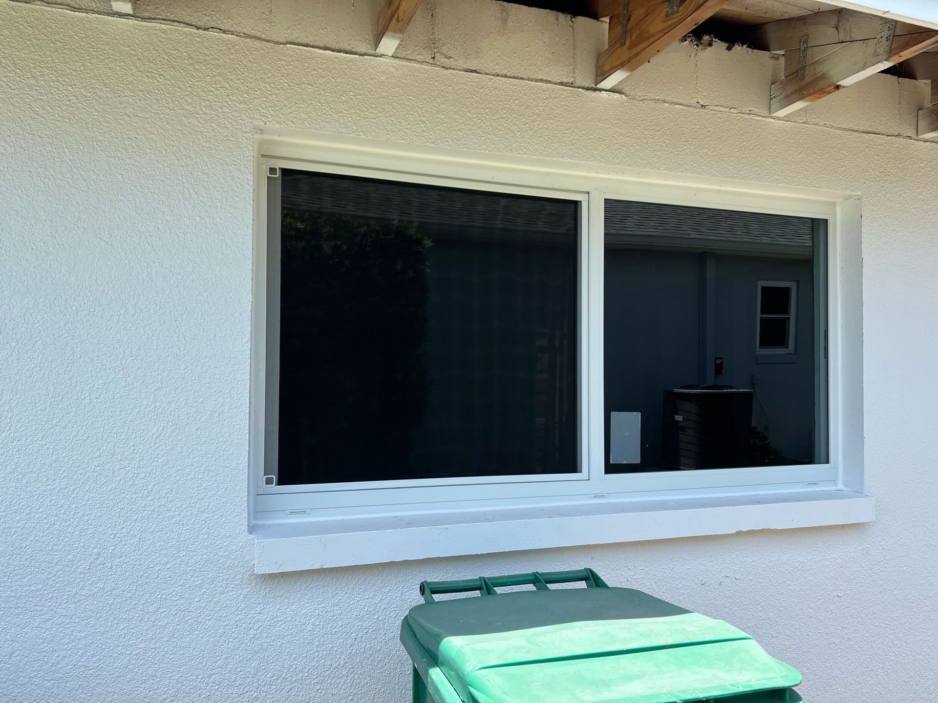 A green trash can is sitting in front of a window on the side of a building.