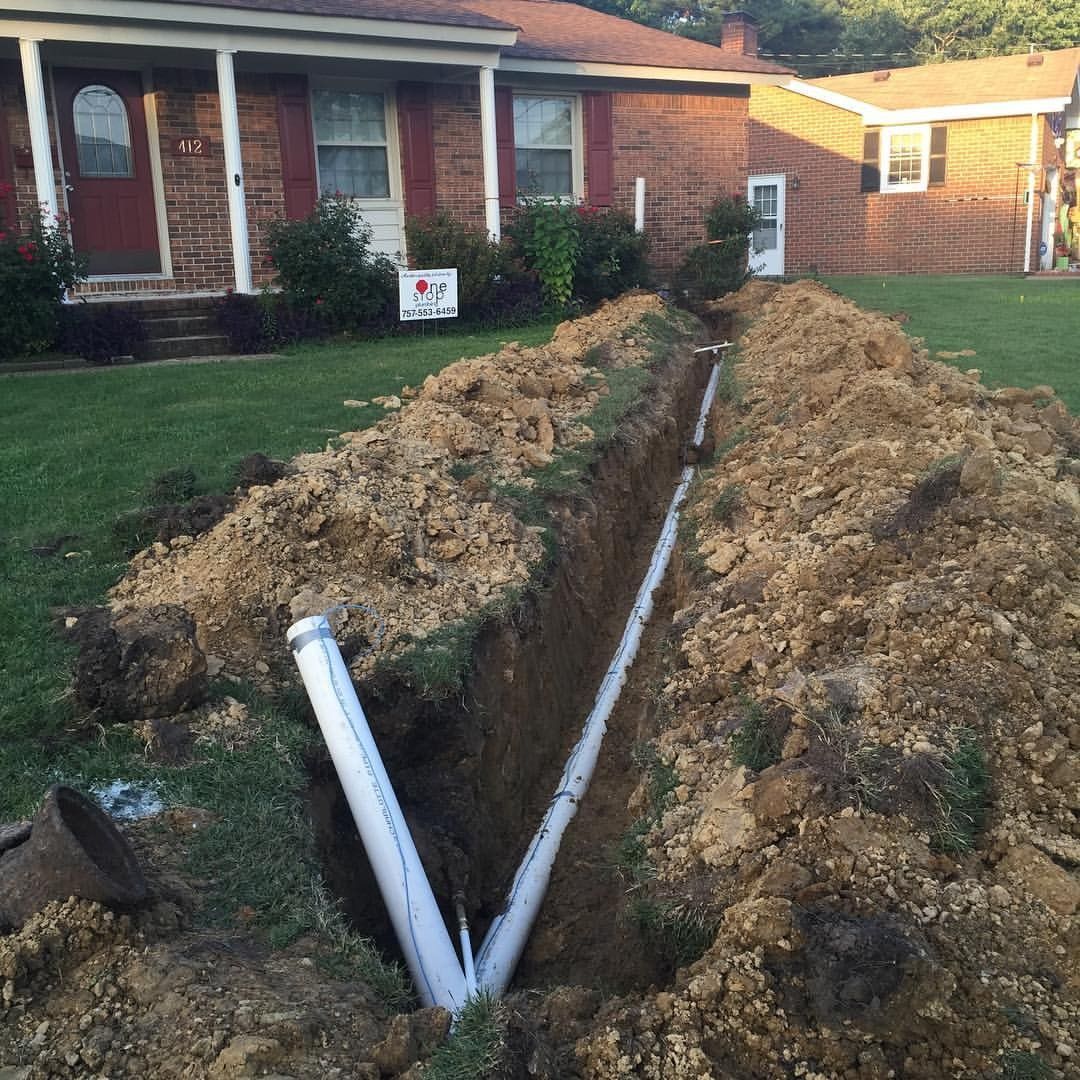 Trenched yard with exposed white pipes running between a brick house and a detached garage.