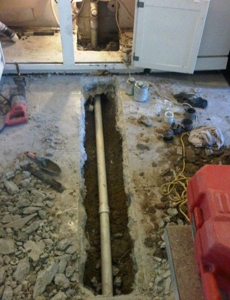 Trench in a concrete floor with exposed white pipe work under open cabinet doors during repair