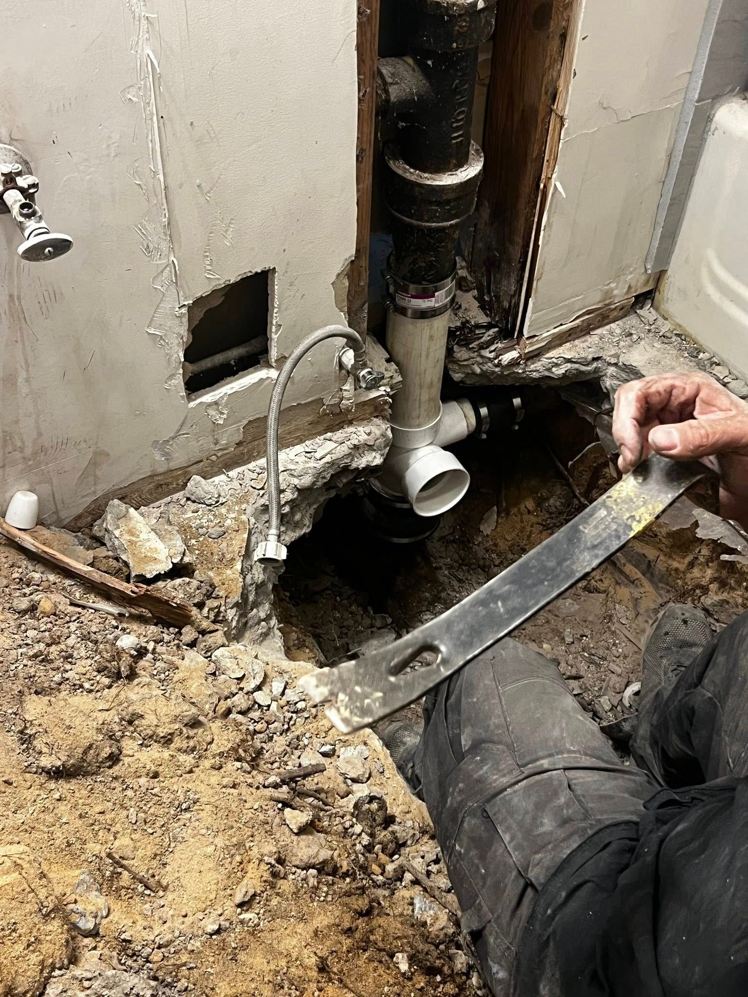 Worker repairing exposed plumbing and broken floor beside a bathroom wall