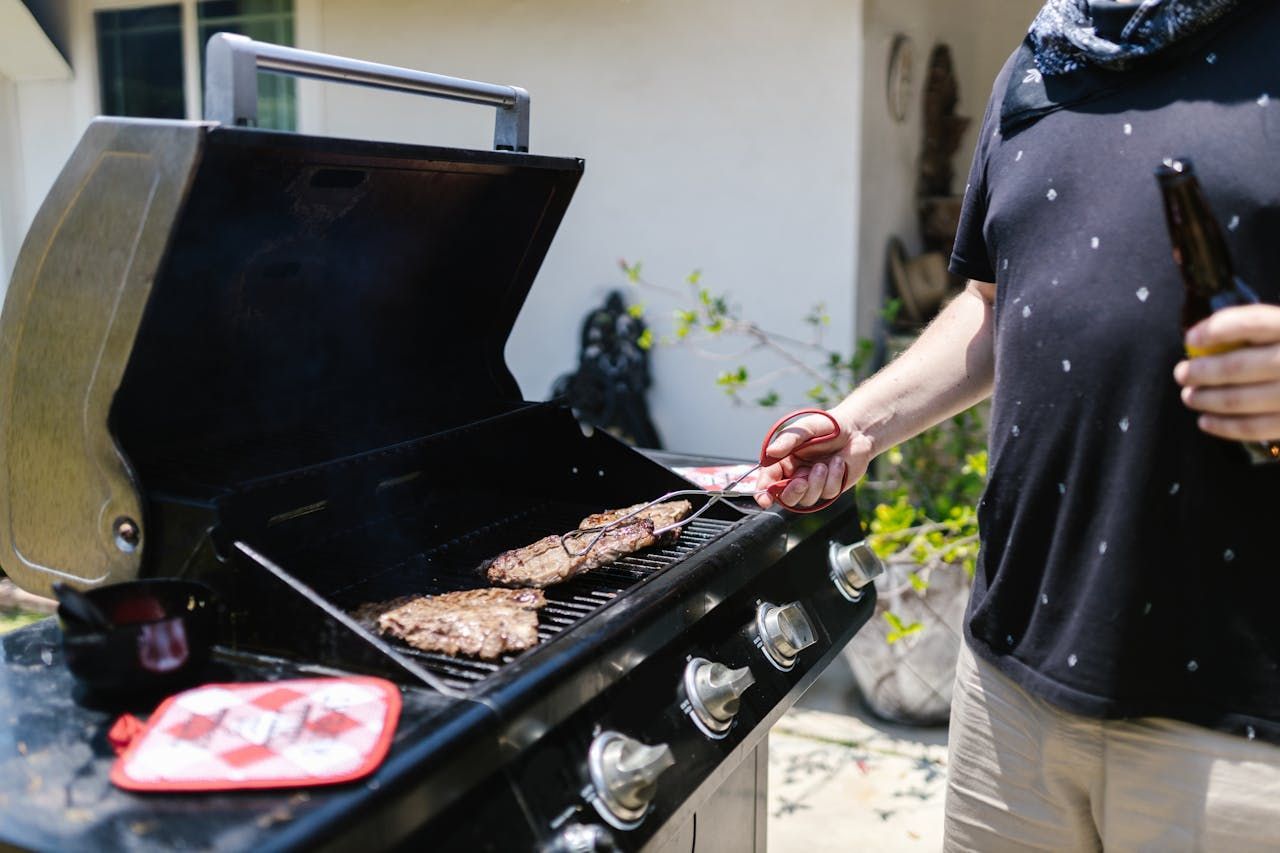 Person grilling meat on a black barbecue outdoors, holding tongs beside the open lid.