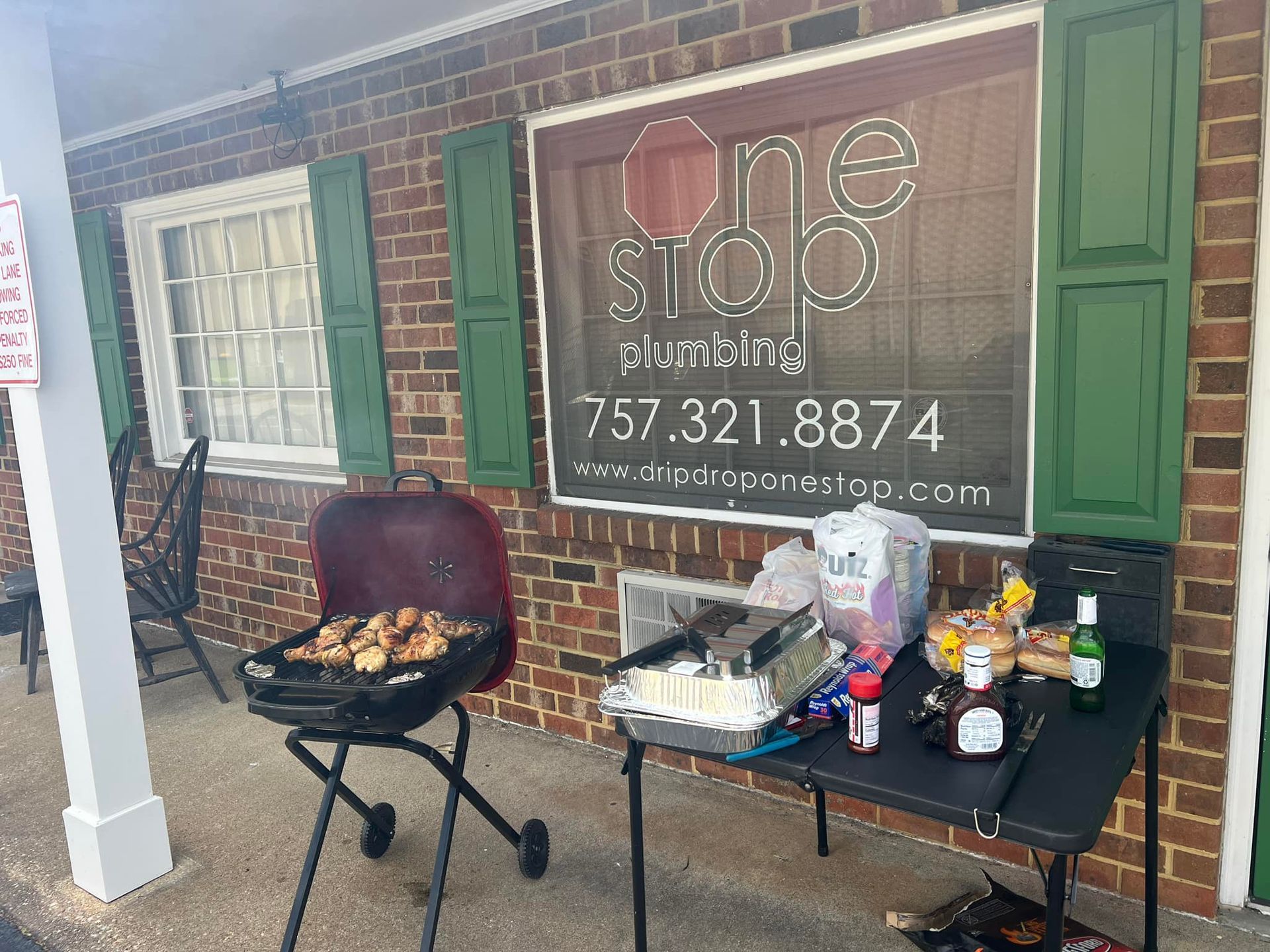 Grilled food on a charcoal grill beside a table with supplies and a banner outside a brick storefront