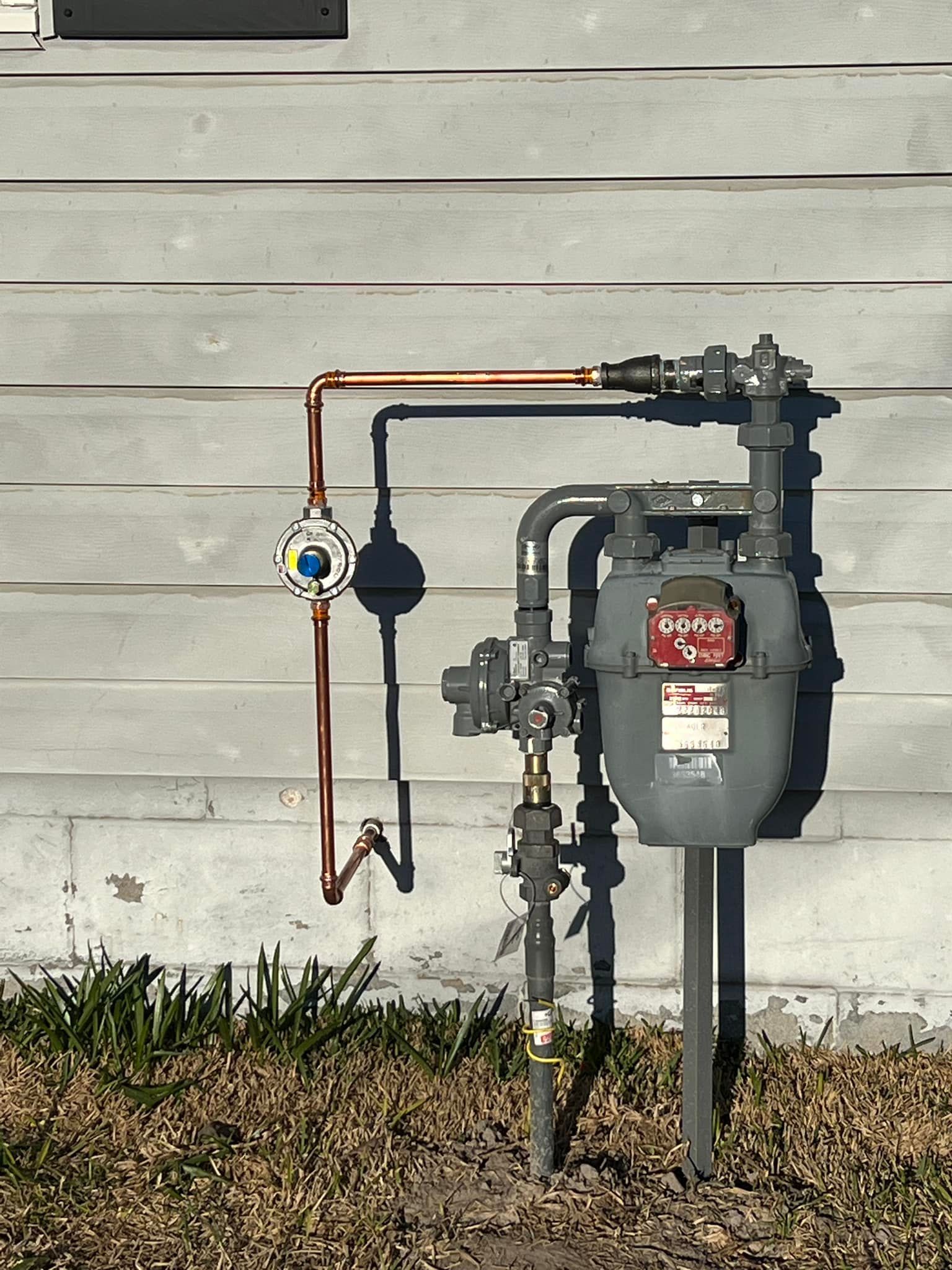Gas meter and copper utility pipes mounted on a house exterior wall with a small lawn below
