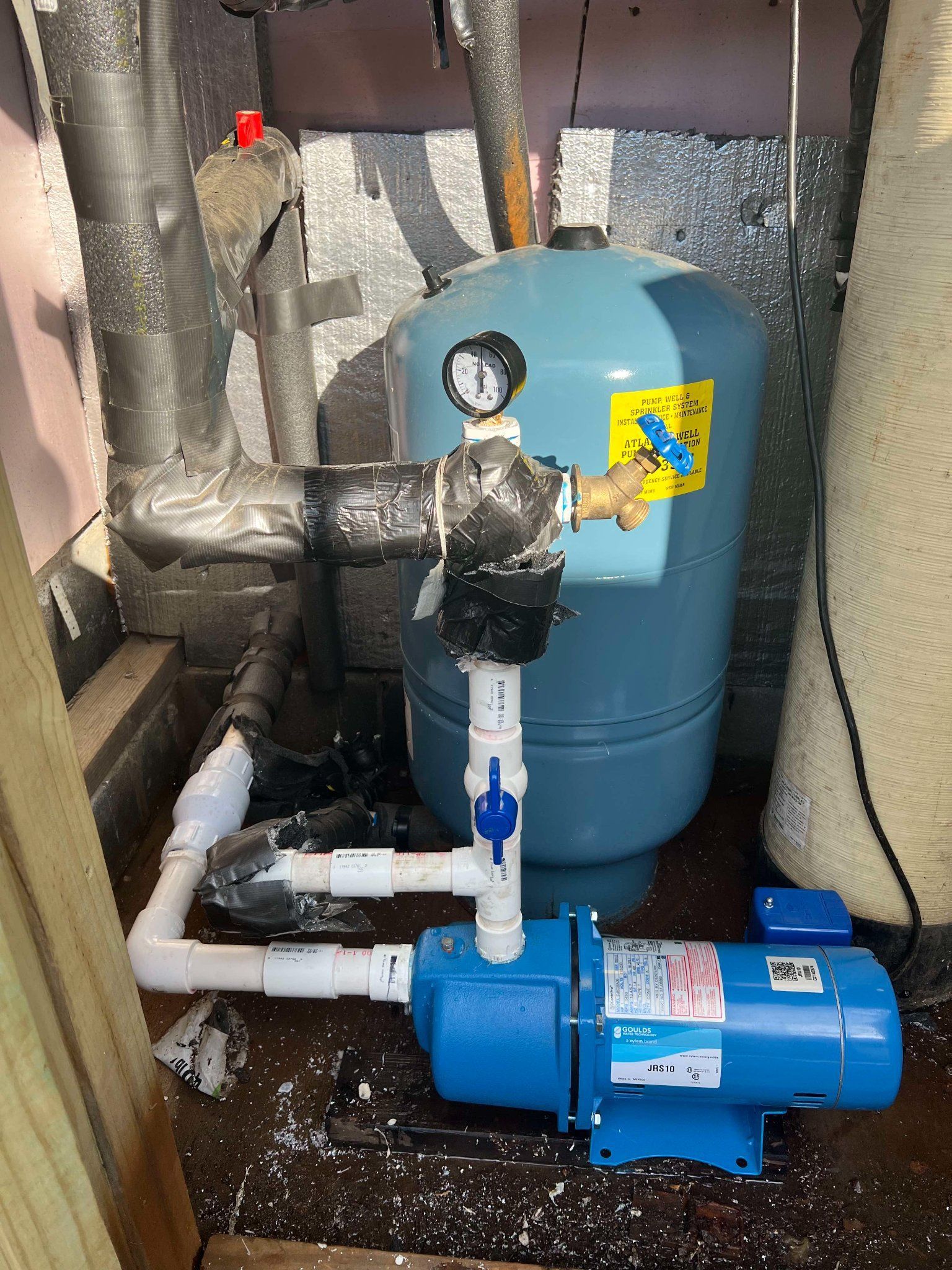 Blue water pump and pressure tank with pipes in a utility room, wrapped with insulation.