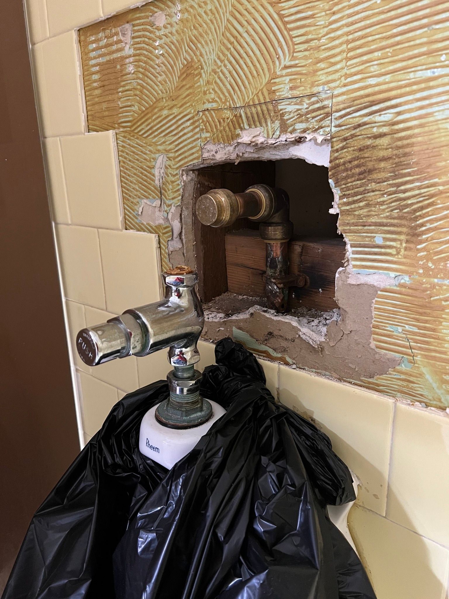 Wall with broken tile exposing plumbing; black trash bag below and a drill on a mounted tool.
