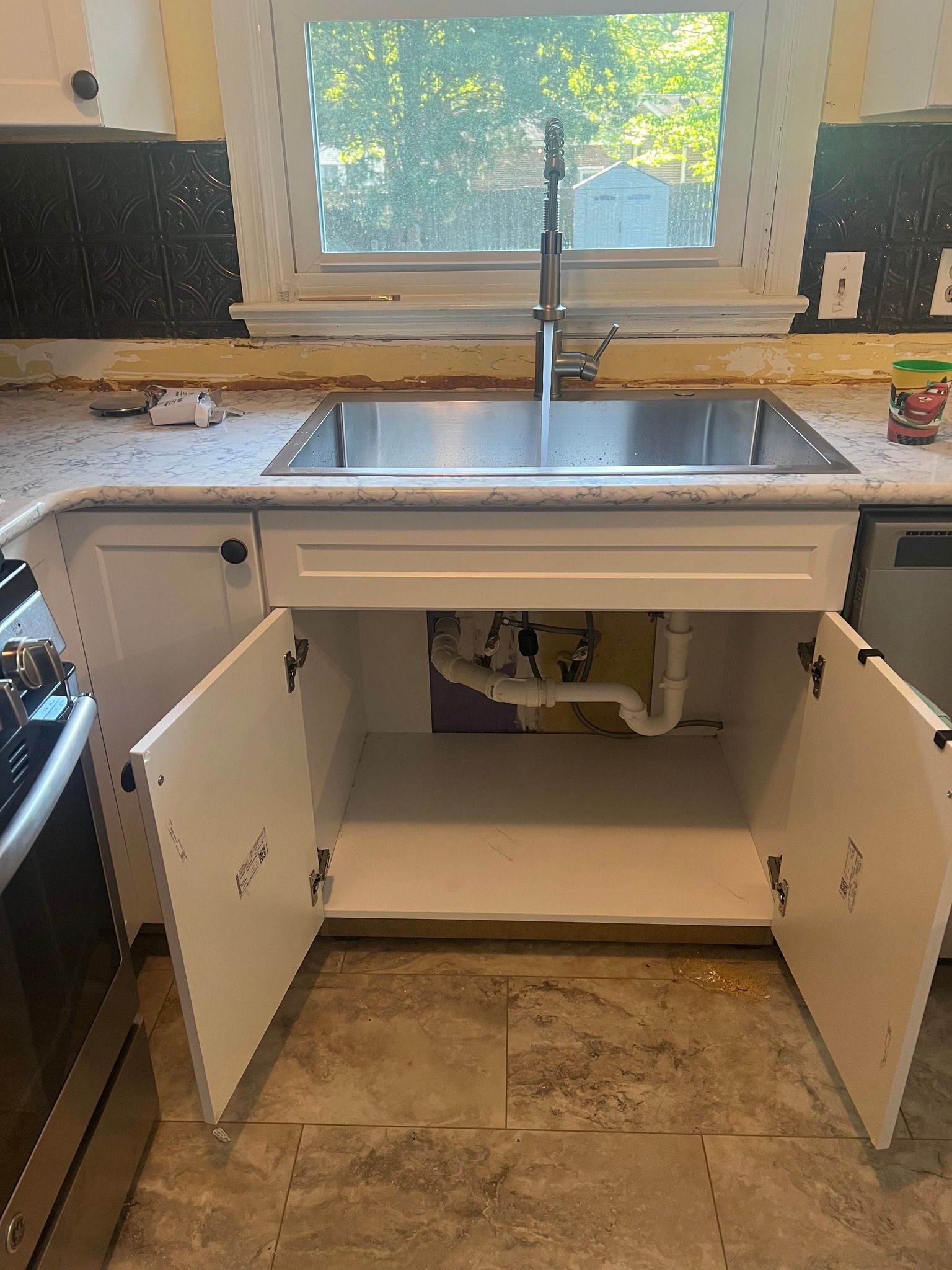 Open kitchen sink cabinet with white doors, stainless sink, and exposed plumbing under a window.