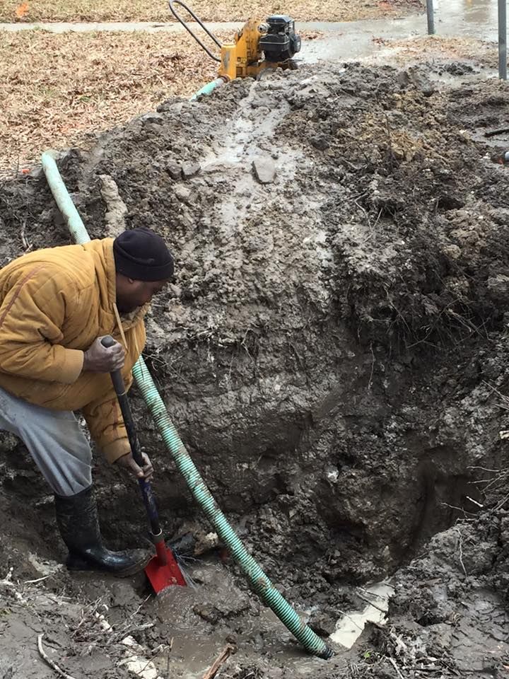 Person digging a muddy trench with a shovel near a large dirt pile outdoors