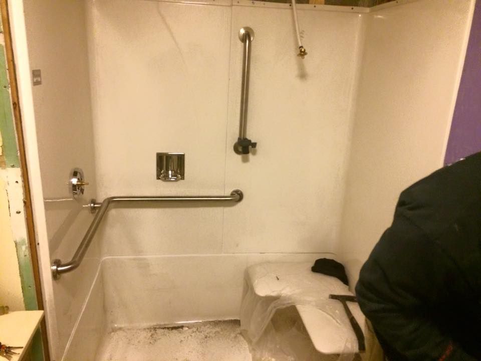 Accessible bathroom with white shower, grab bars, and a shower chair; person partially visible at right.
