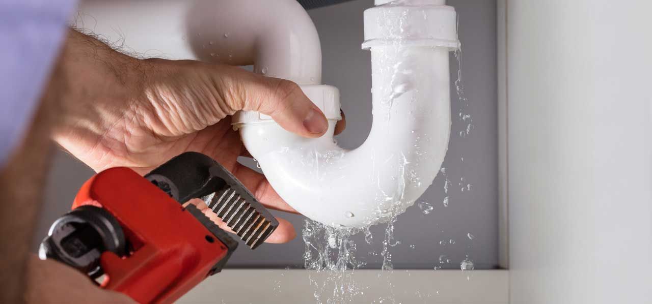 Hand using a red pipe wrench to tighten a white PVC drain pipe under a sink, with water spraying out