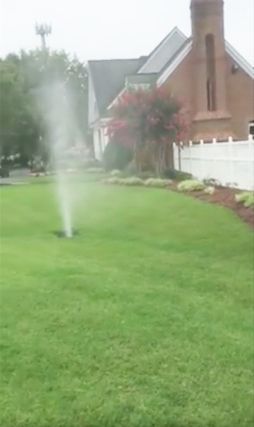 Lawn sprinkler spraying water beside a brick house and white fence on a green suburban yard