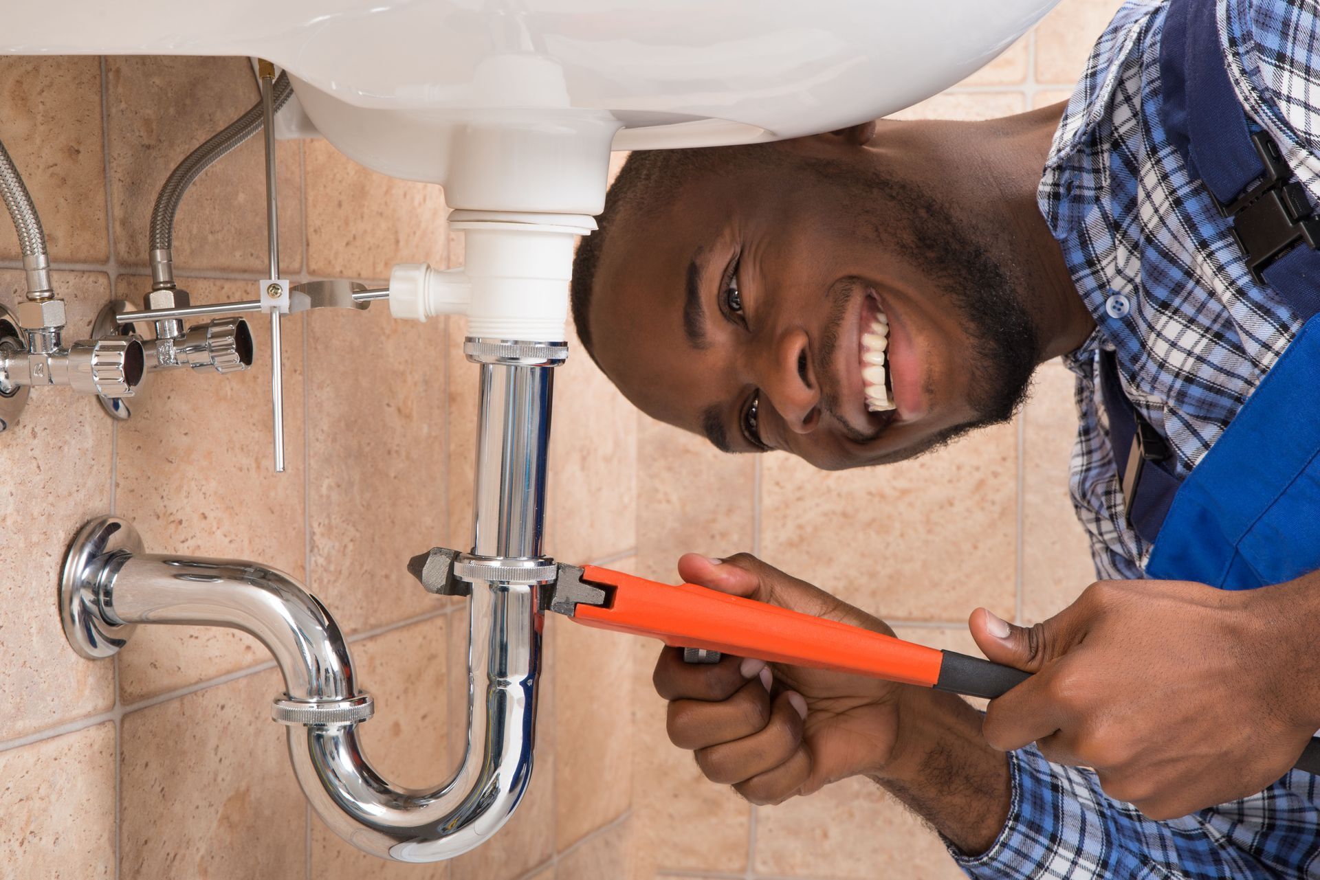 Plumber tightening a chrome pipe under a sink with a red wrench, smiling in a bathroom.