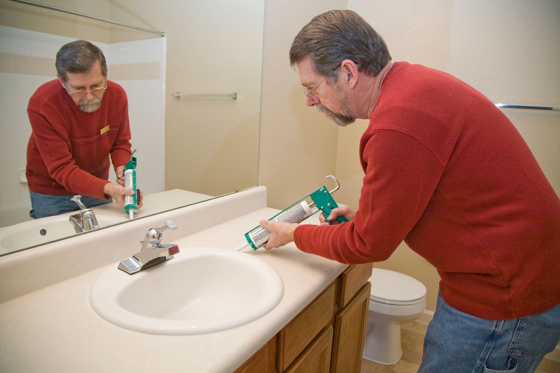 Man cleaning a bathroom sink with a green spray bottle, reflected in the mirror