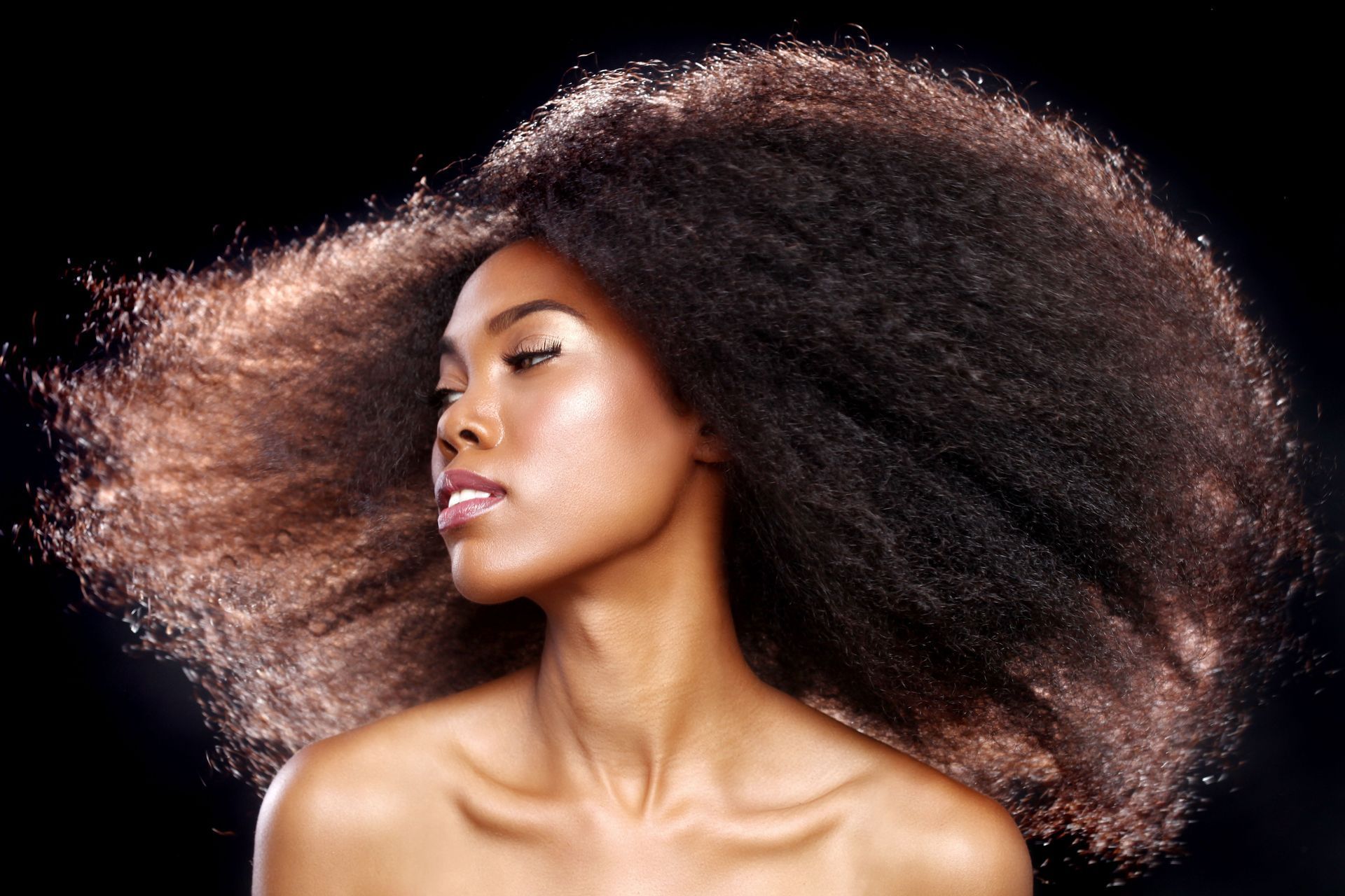 Woman with dark, voluminous afro, head turned to the side, against a black background.