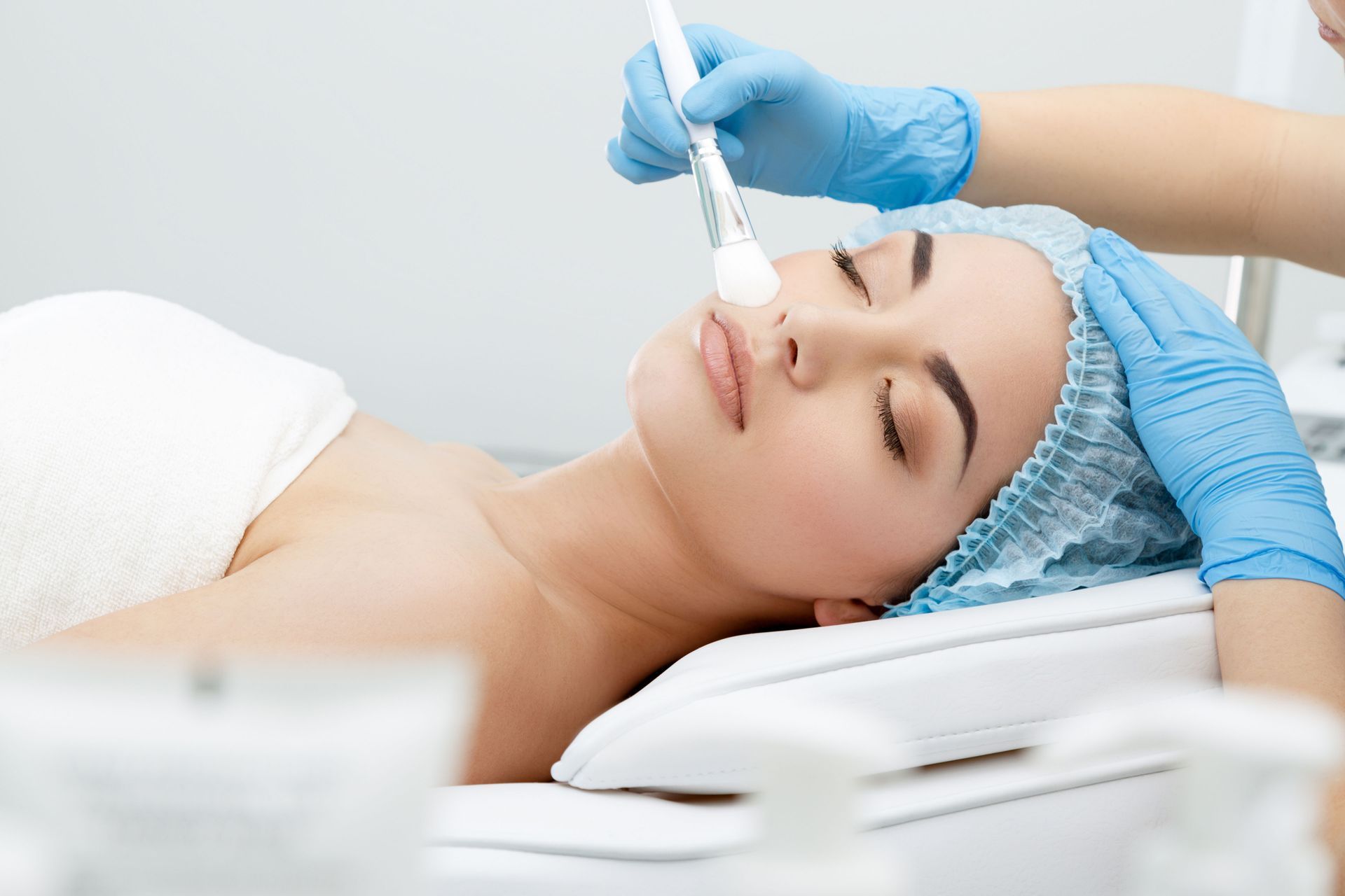 Woman receiving facial treatment with a brush in a spa. Blue gloves, cap, and white towels.