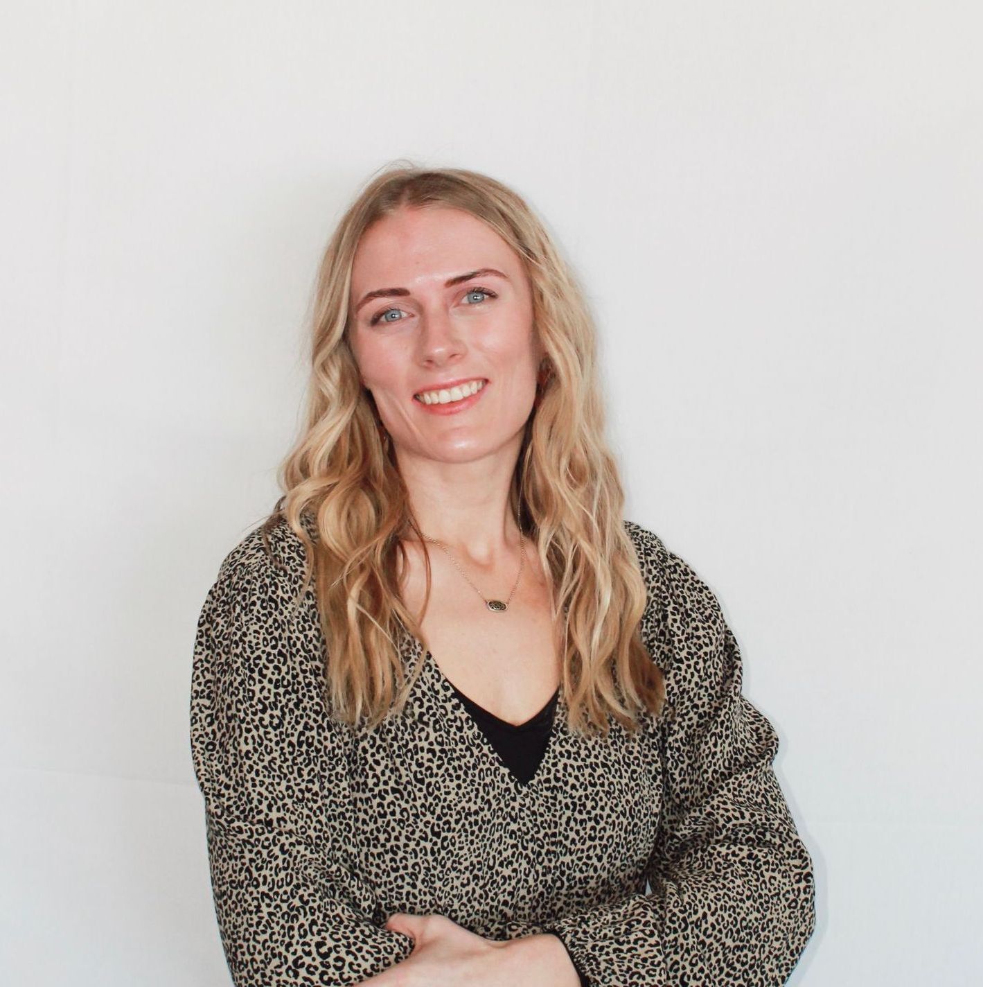 Lilee Cox with wavy hair smiles, wearing a black v-neck and animal print top, in front of a white wall.