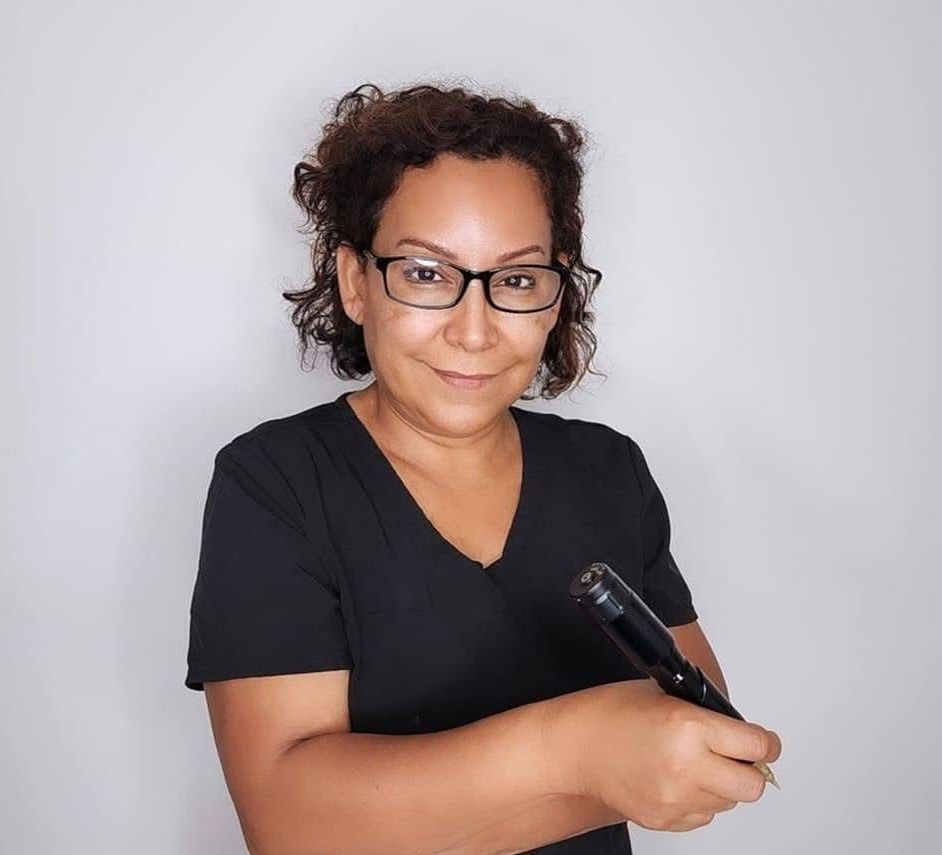 Martha Turvey with glasses, black shirt, holding a black object, smiling against a gray background.