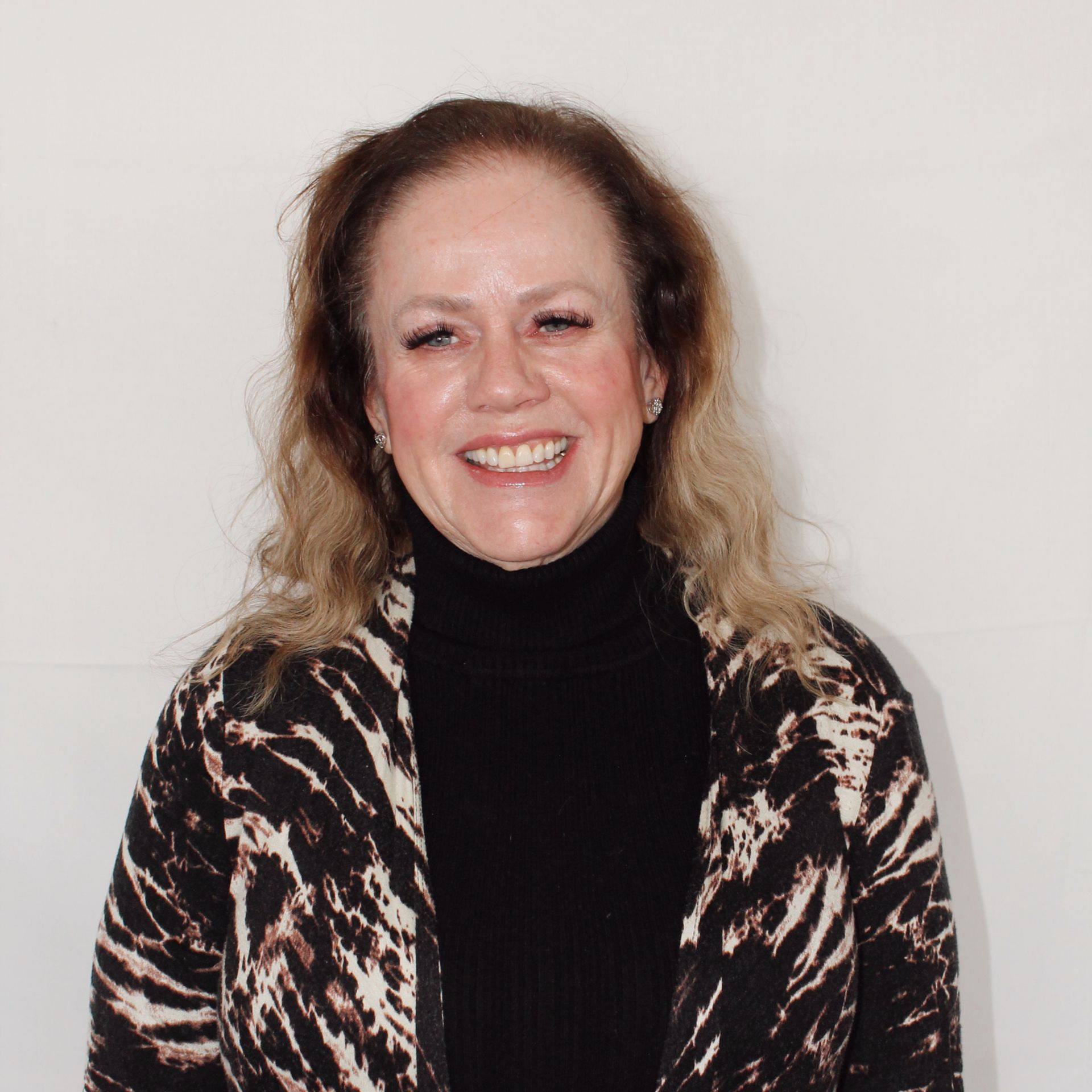 Michaelle Holland with curly blonde hair wearing a black turtleneck and animal print coat smiles in front of a white wall.