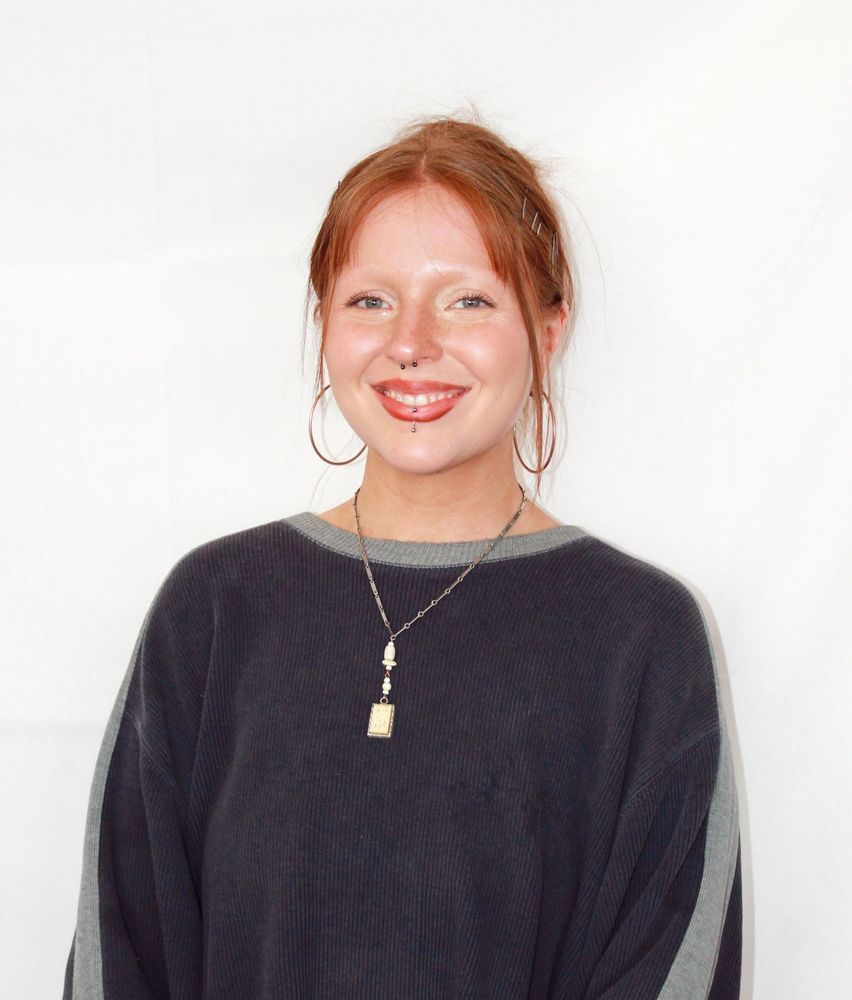 Regan Corby with red hair, nose ring, hoop earrings, smiling, wearing a gray sweater and necklace.