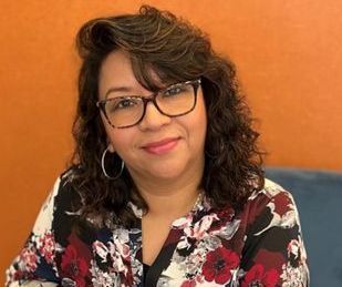 Sara Alvarado with glasses, dark hair, floral shirt, smiling, orange background.