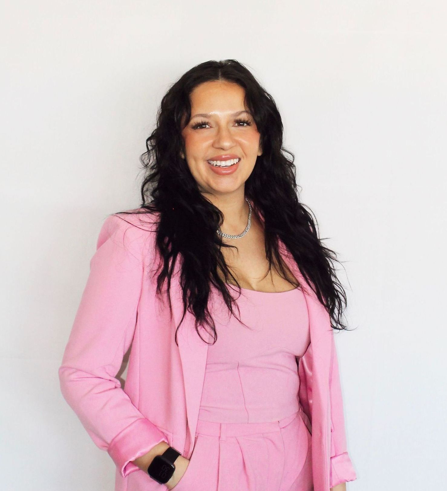Serene Stricklin with long black hair wearing a pink suit, smiling against a white background.